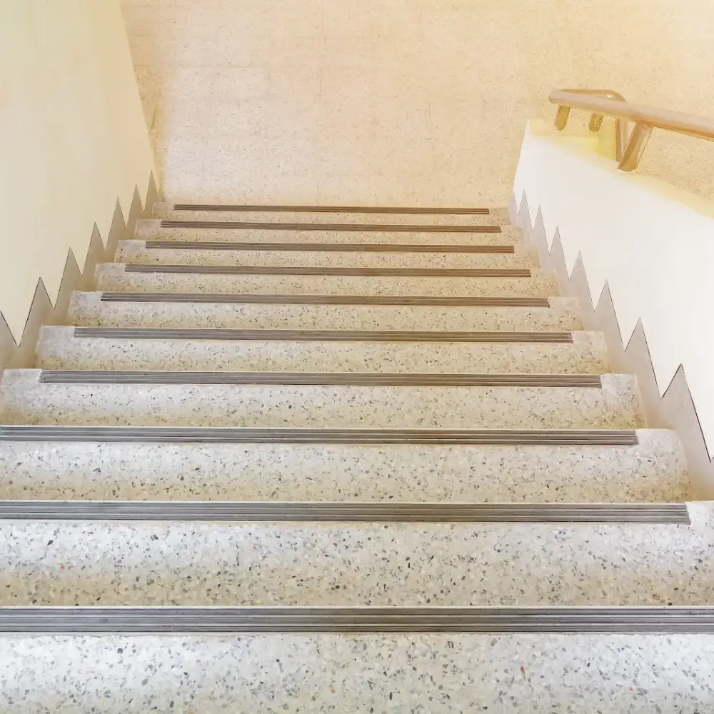 Indoor staircase with terrazzo steps and metal anti-slip strips, bordered by white walls and a handrail.