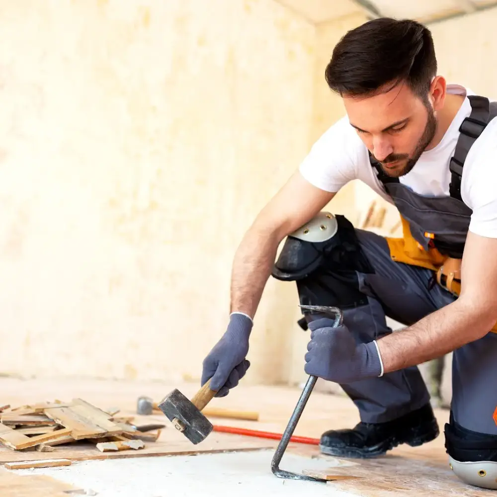 Construction worker removing old flooring with a hammer and pry bar while wearing protective gloves and knee pads.