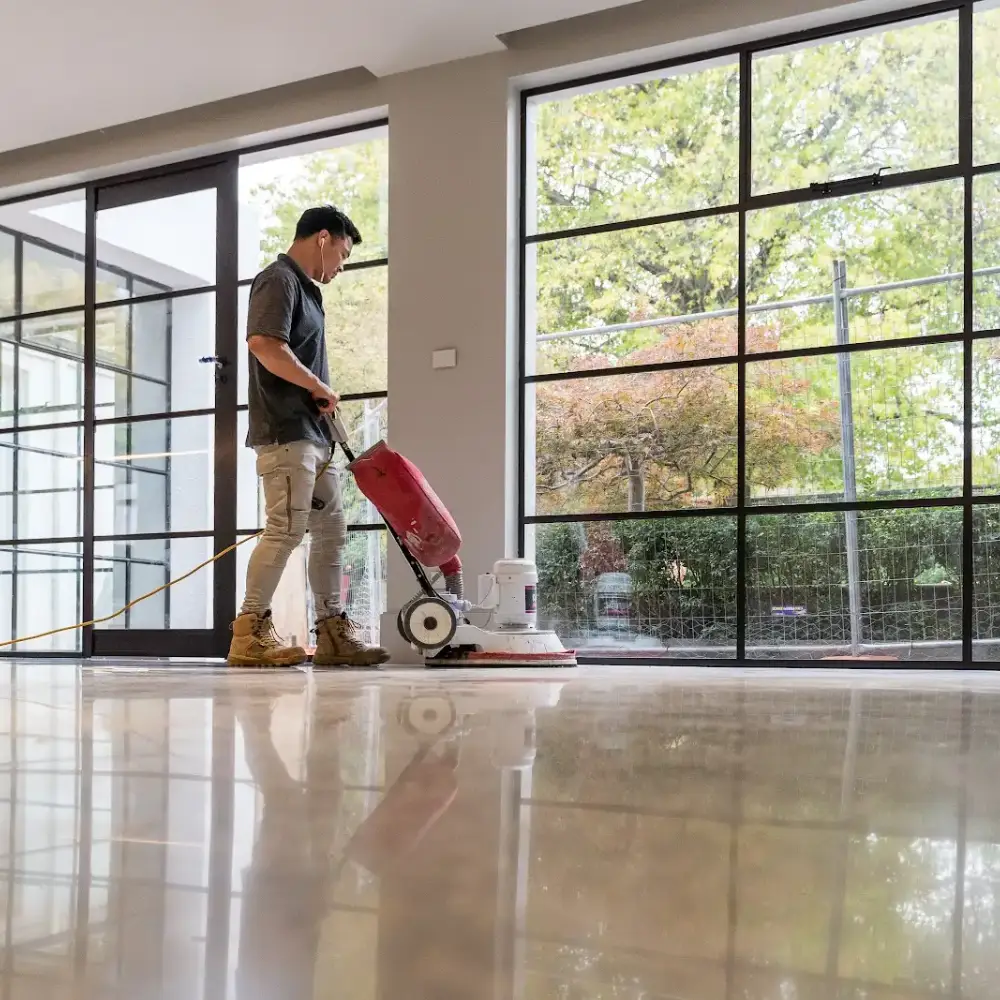 Worker polishing a concrete floor with a floor buffer in a modern room with large glass windows.