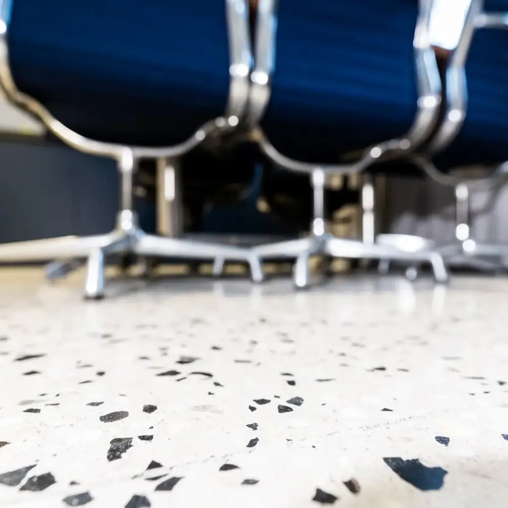 Close-up of a light-coloured polished concrete floor with black aggregate, beneath blue office chairs on chrome bases.