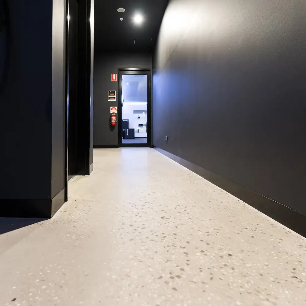 Light-coloured polished concrete floor in a modern hallway with dark walls leading to a brightly lit room.