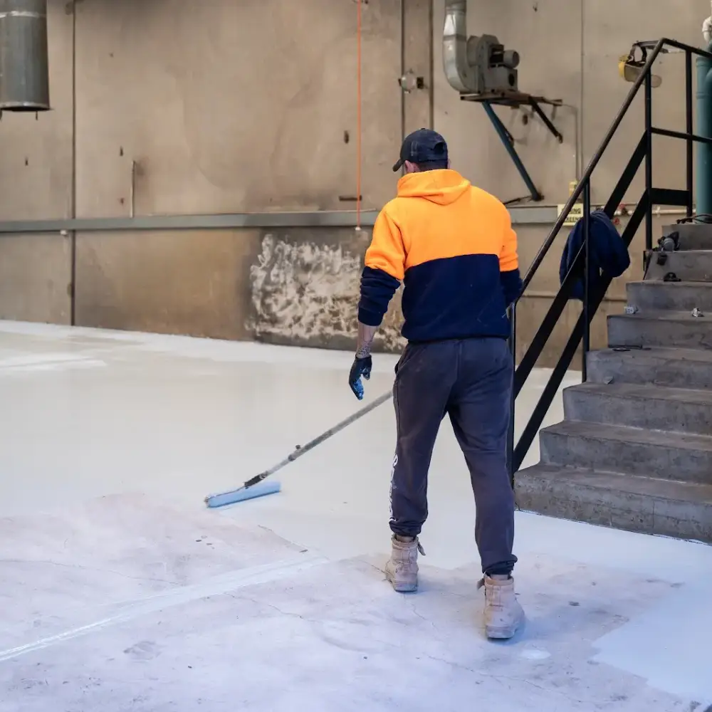 Worker in a high-vis orange hoodie applying epoxy coating with a roller on a large warehouse concrete floor near a staircase.