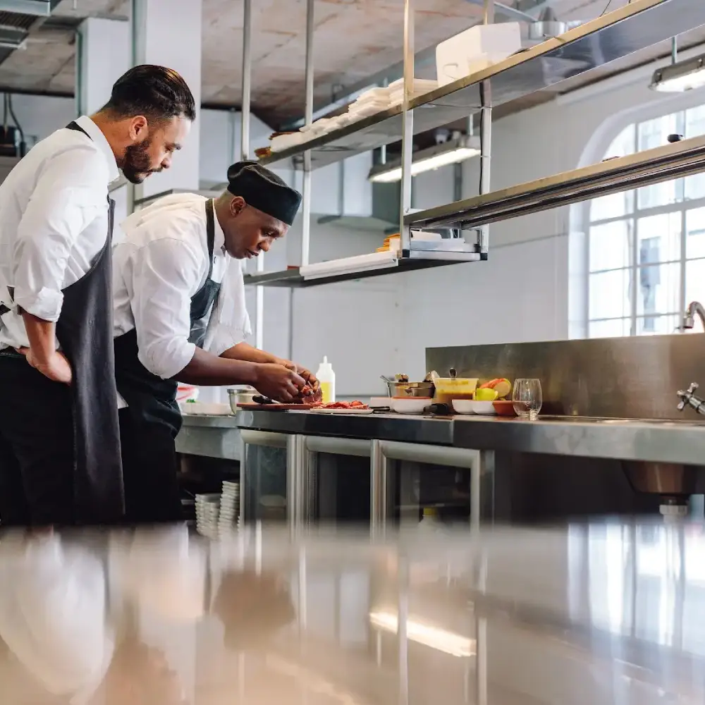 Two chefs working in a commercial stainless steel kitchen, preparing food at a counter with fresh ingredients and utensils.