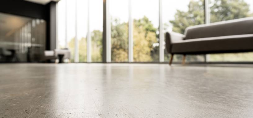 Low-angle shot of a smooth, polished concrete floor in a bright, modern office space with large windows and blurred sofas.