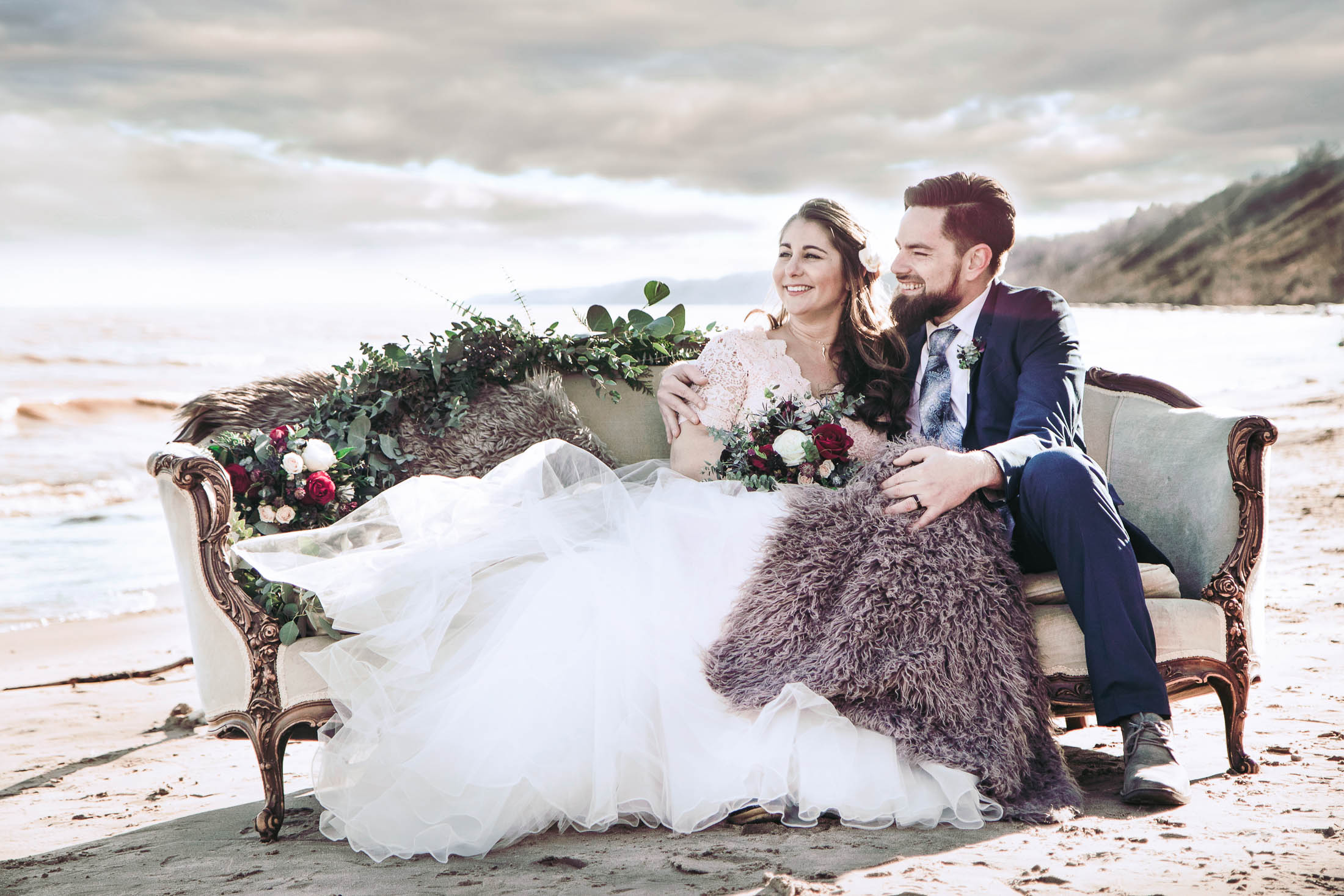 Bride and groom on antique sofa on beach with flowers and greens and furry blankets