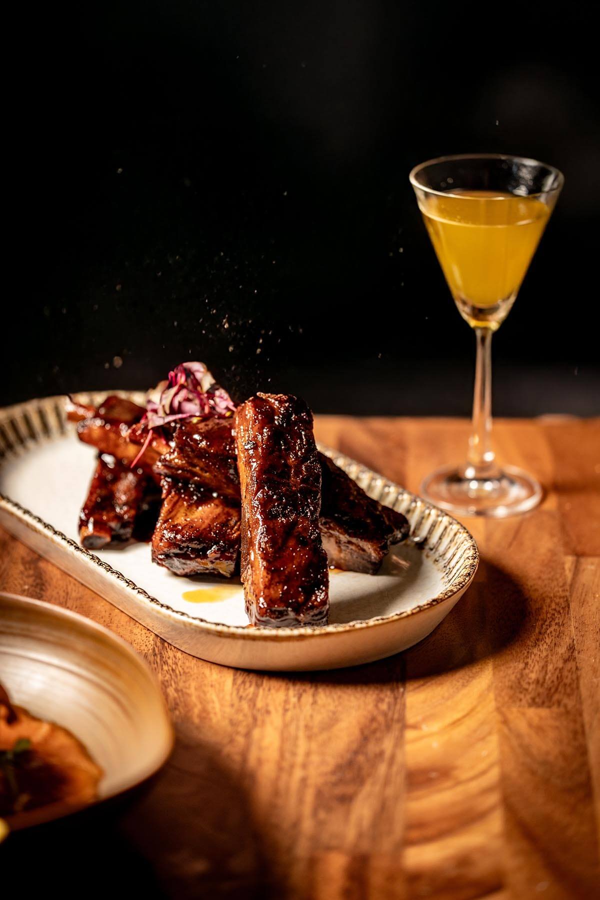 Plate of glazed barbecue ribs garnished with microgreens on a wooden table with a cocktail glass of orange drink.