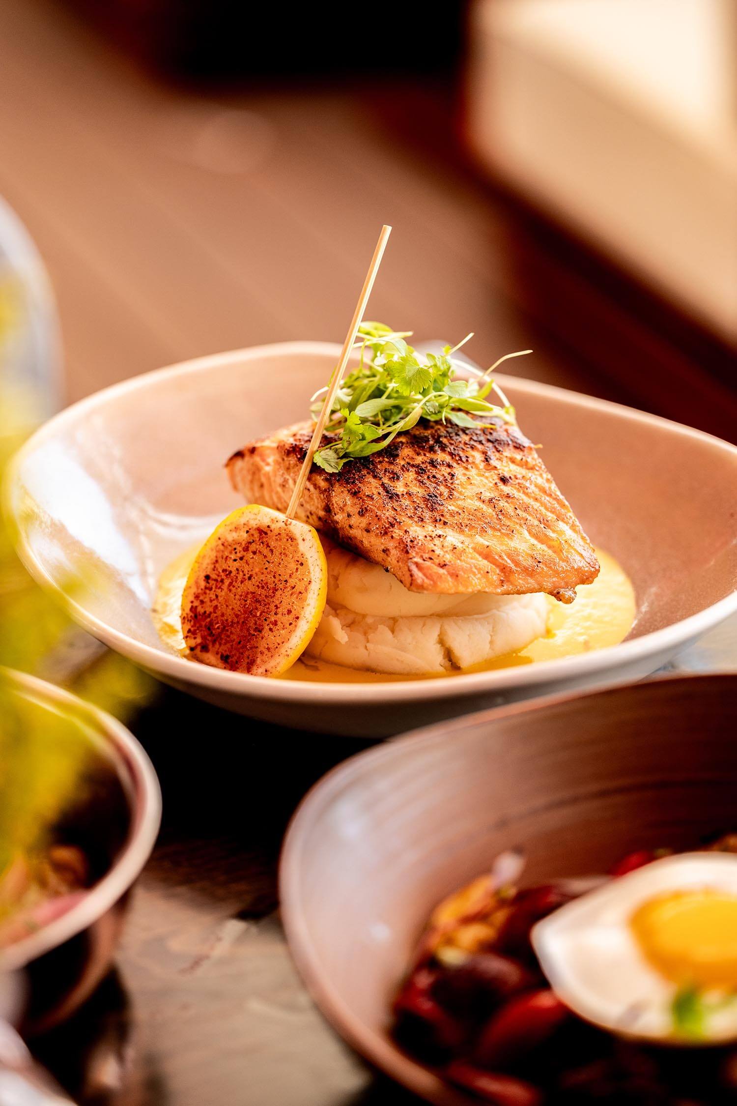 Grilled salmon fillet on mashed potatoes, garnished with microgreens and a grilled lemon half on a beige plate.