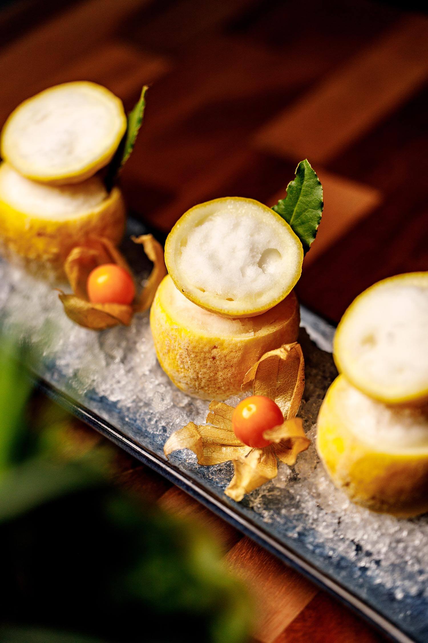 Three yellow dessert cups filled with white foam, garnished with green leaves and orange physalis berries on a bed of crushed ice.