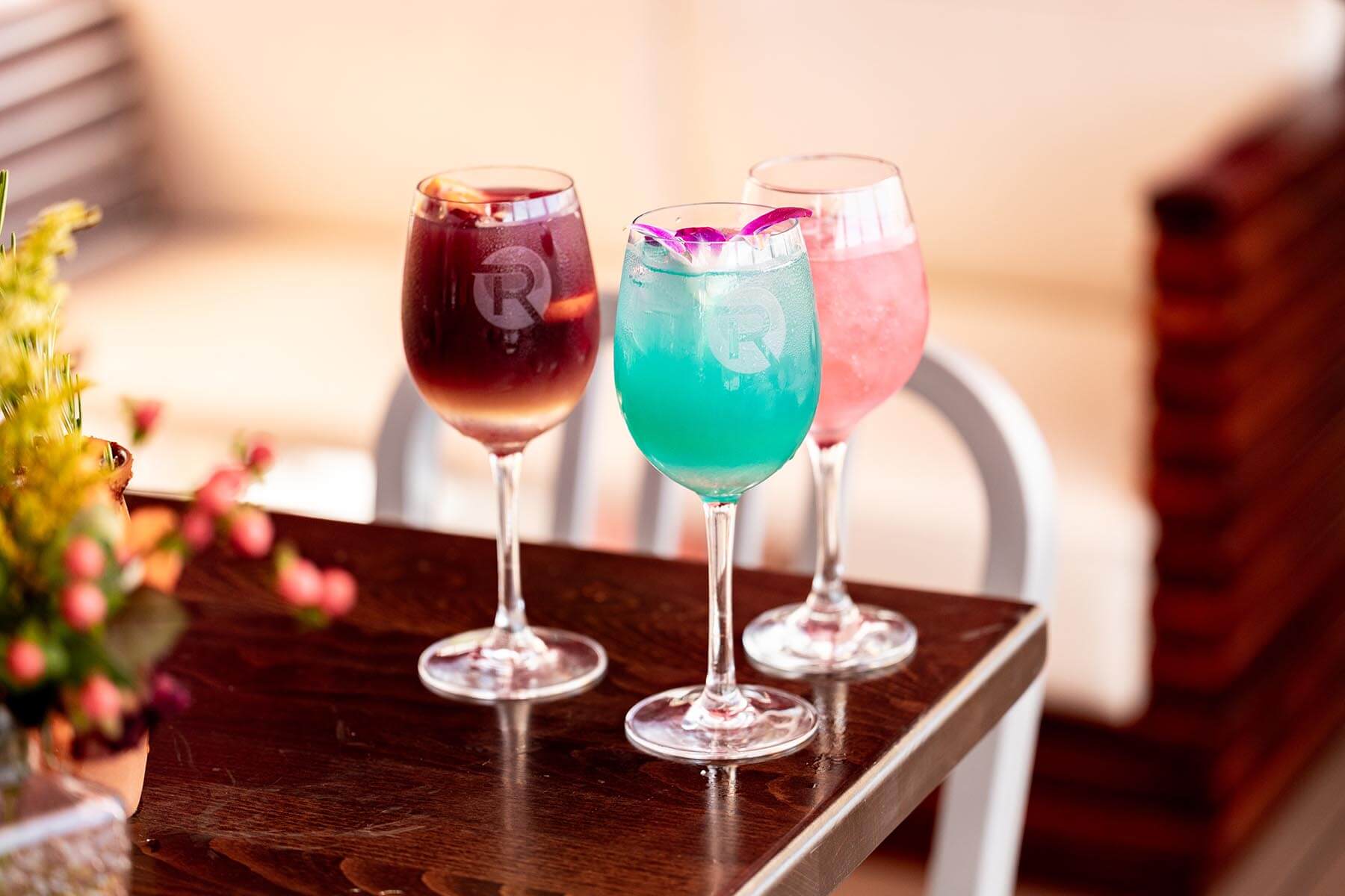 Three colorful cocktails in wine glasses on a wooden table, one red, one turquoise with a purple flower garnish, and one light pink.