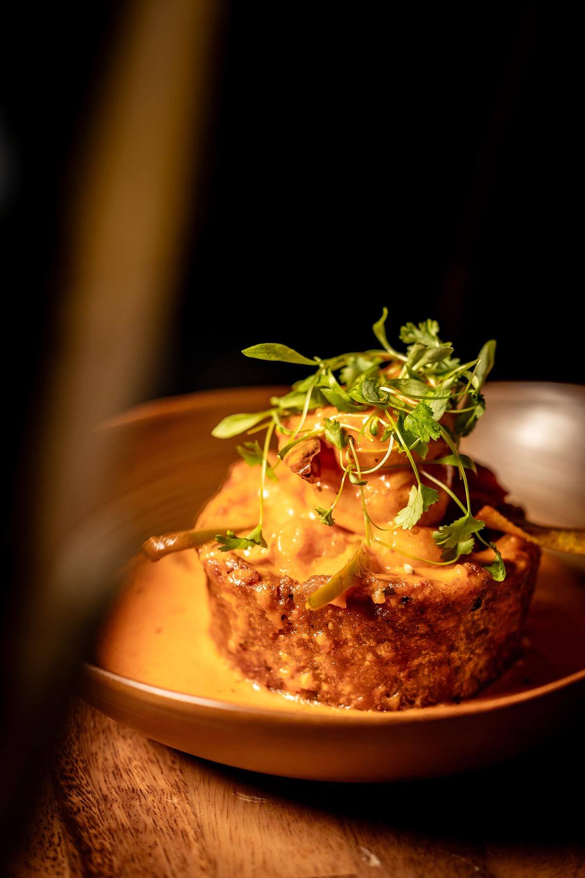 Close-up of a crispy, round savory dish topped with orange sauce and fresh green herbs on a brown plate.