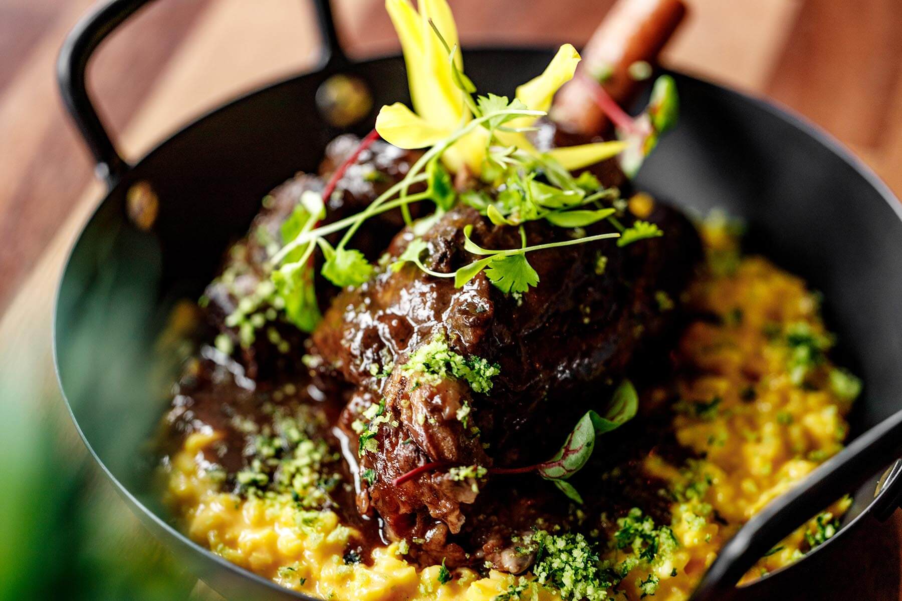 Braised meat shank garnished with herbs served on creamy yellow risotto in a black pan.