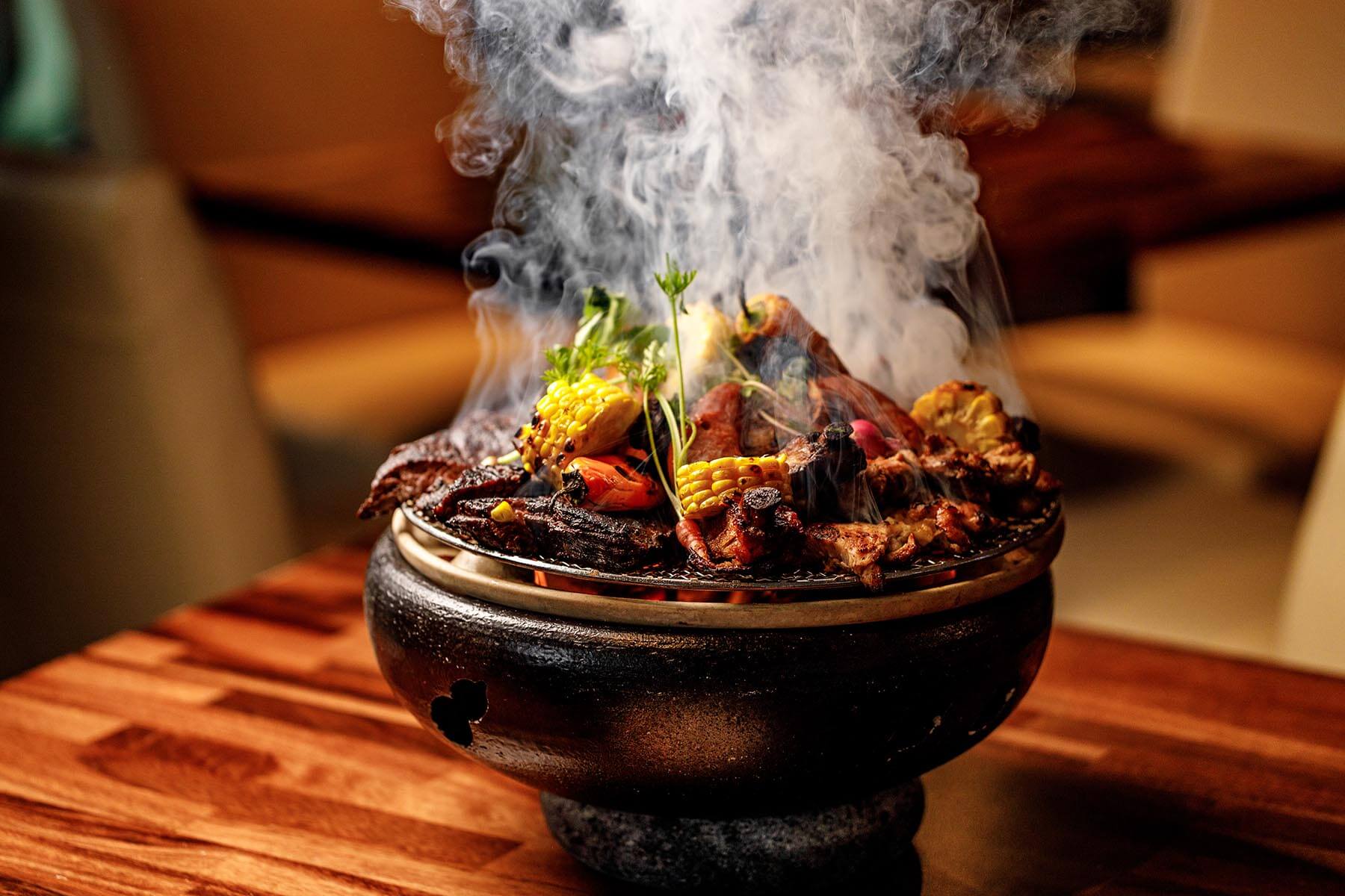 Smoky charcoal grill with assorted grilled meats, corn, and vegetables on a wooden table.