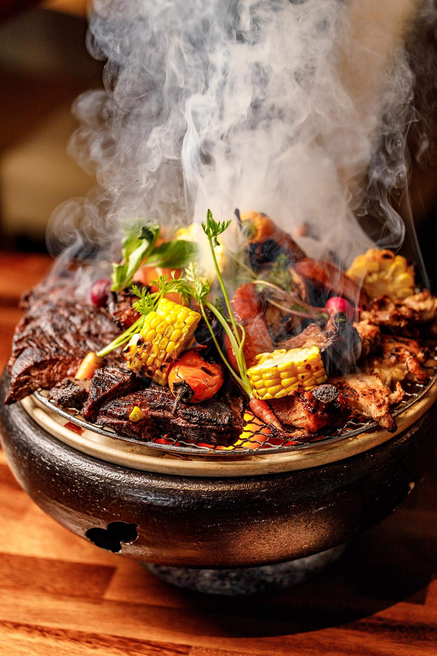 Smoke rising from a traditional charcoal grill loaded with grilled meat, corn, carrots, and vegetables.