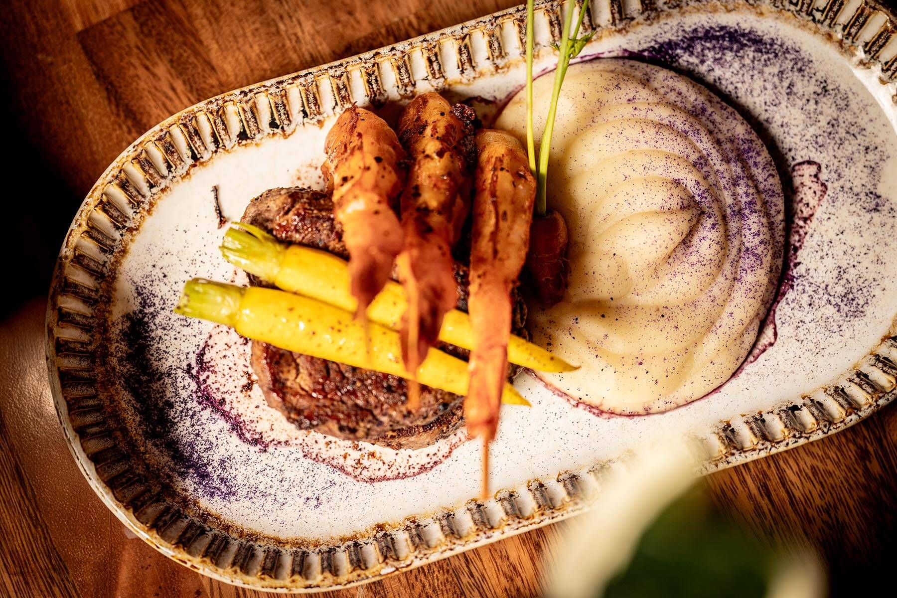 Grilled steak topped with yellow and orange carrots served with a swirl of mashed potatoes sprinkled with purple seasoning on a rustic oval plate.