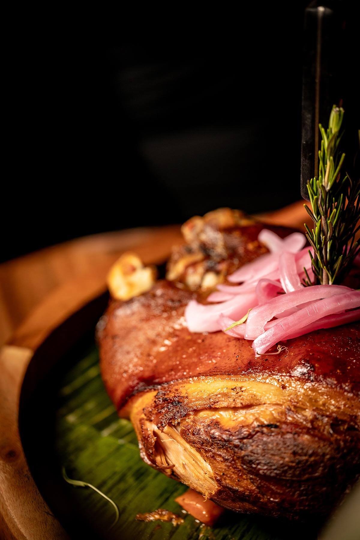 Close-up of a roasted pork dish garnished with pickled red onions and a sprig of rosemary on a green leaf in a wooden bowl.