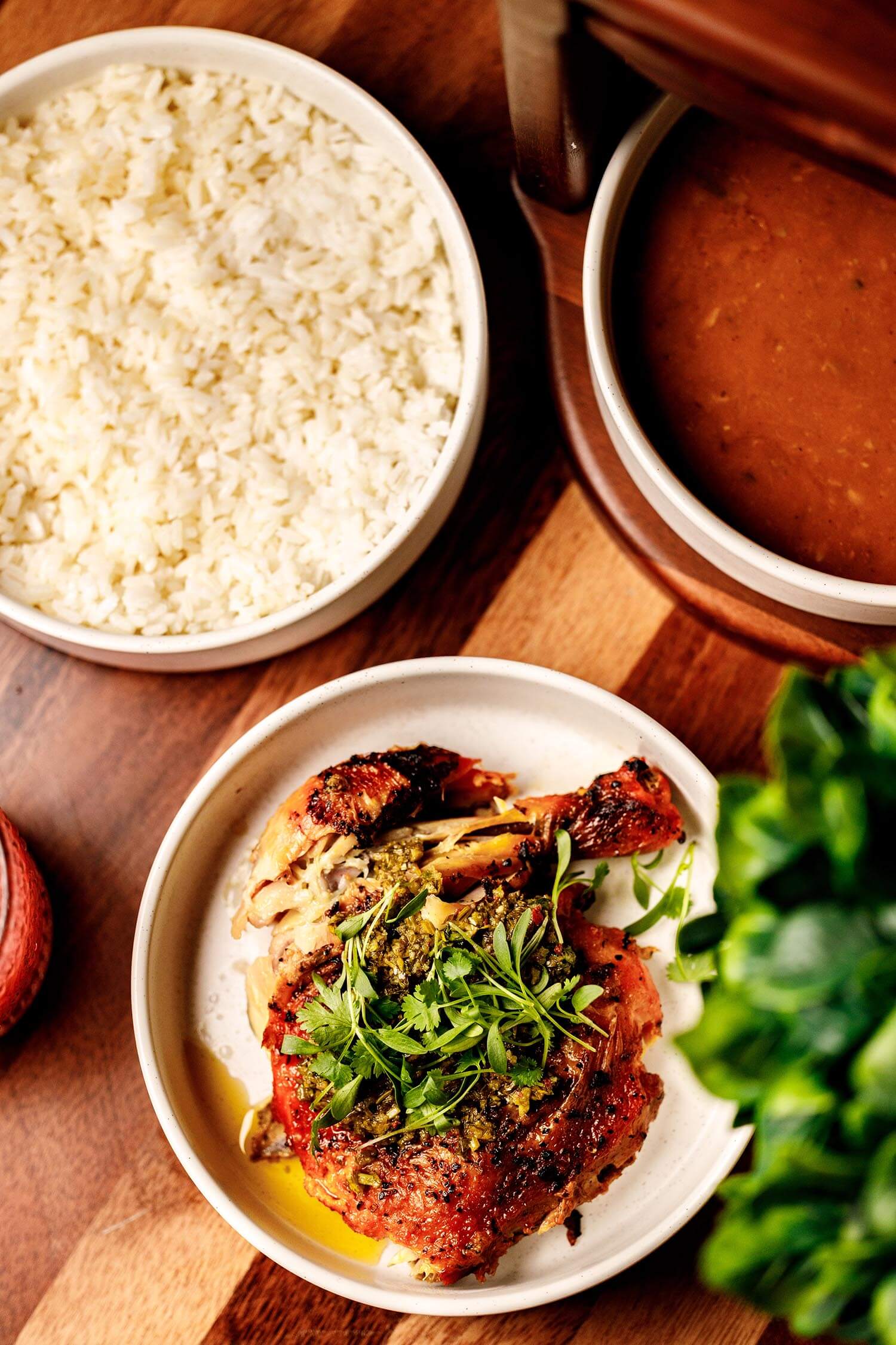 Plate with roasted chicken topped with herbs, bowl of white rice, and bowl of brown sauce on a wooden table.
