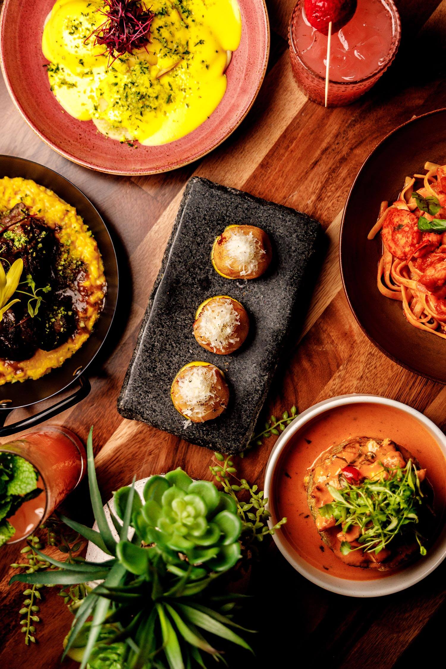 Assorted gourmet dishes including ravioli with yellow sauce, meat over creamy polenta, three small stuffed potatoes with cheese, seafood pasta, and a dish with sauce and greens, accompanied by two cocktails on a wooden table with green plants.