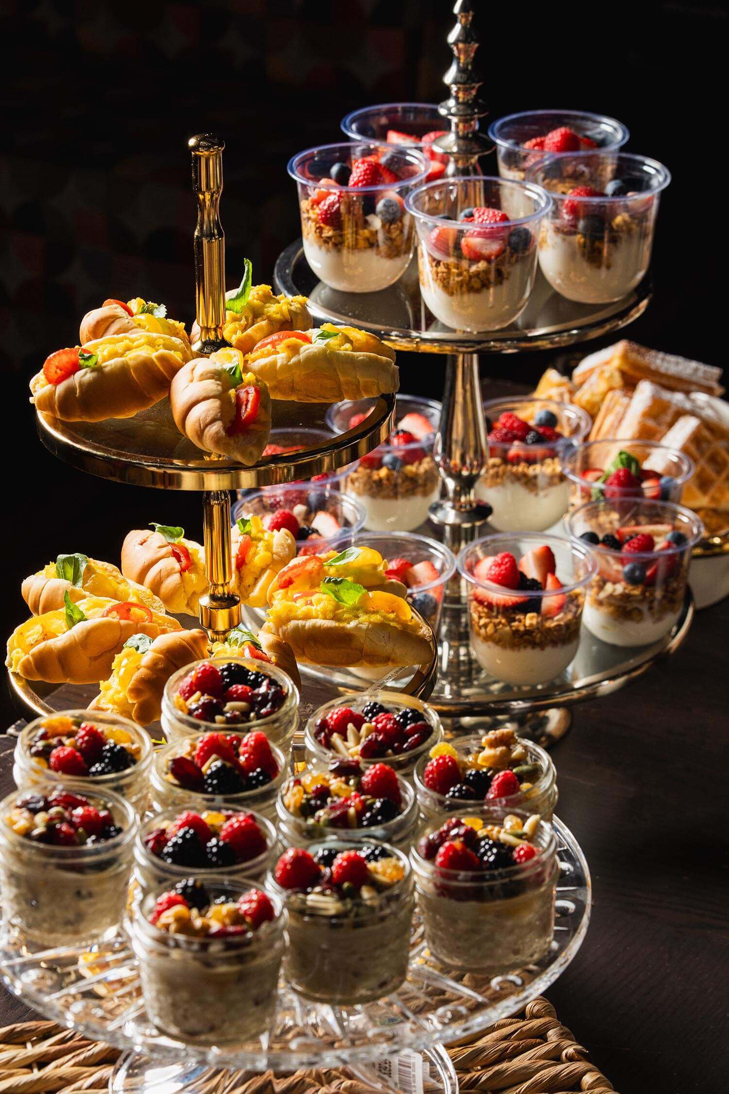 Three-tiered serving stand with assorted breakfast pastries, fruit parfait cups with granola and berries, and jars of oatmeal topped with mixed berries and nuts.