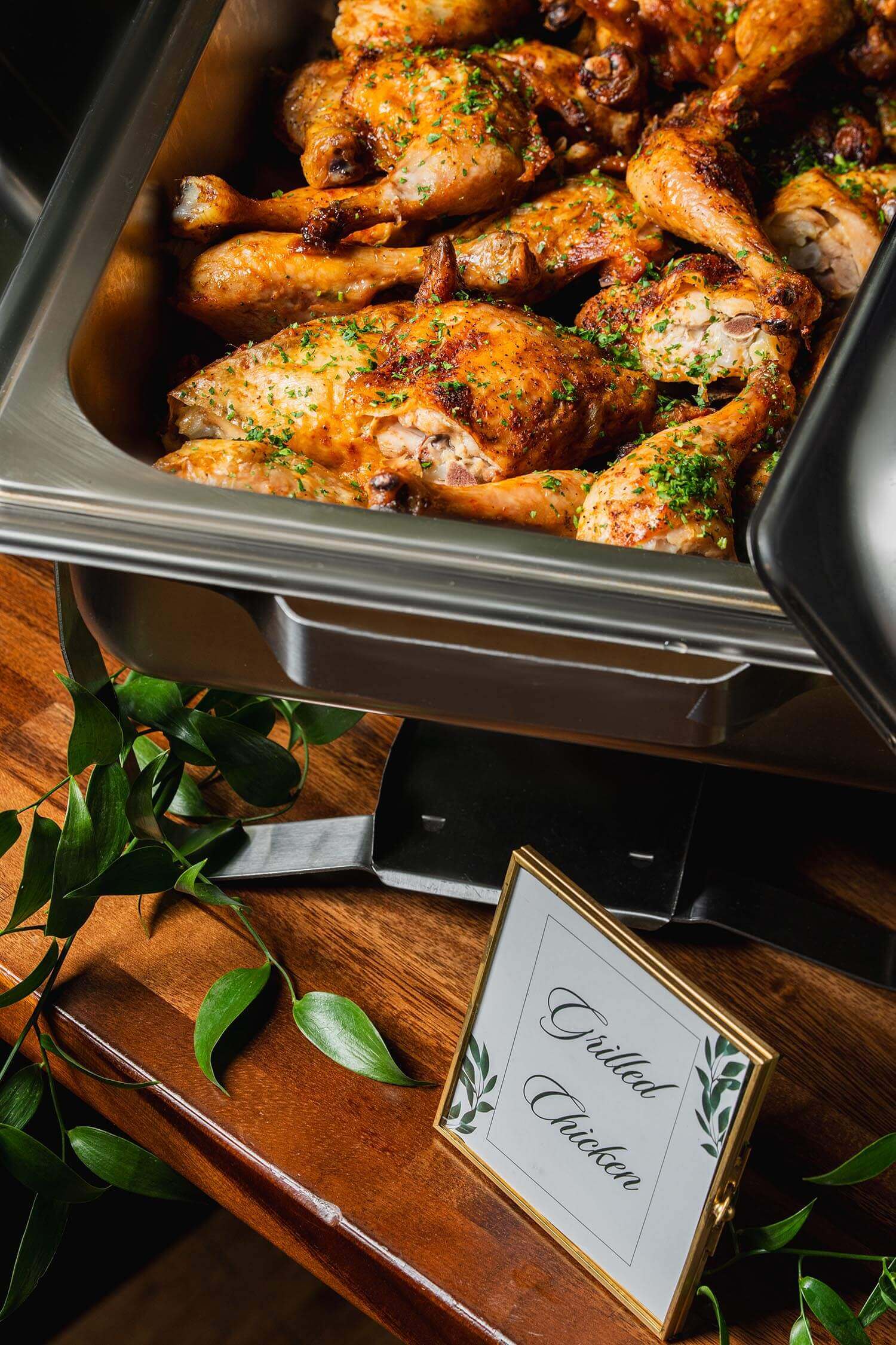 Chafing dish filled with grilled chicken pieces garnished with herbs on a wooden table with a sign reading 'Grilled Chicken.'