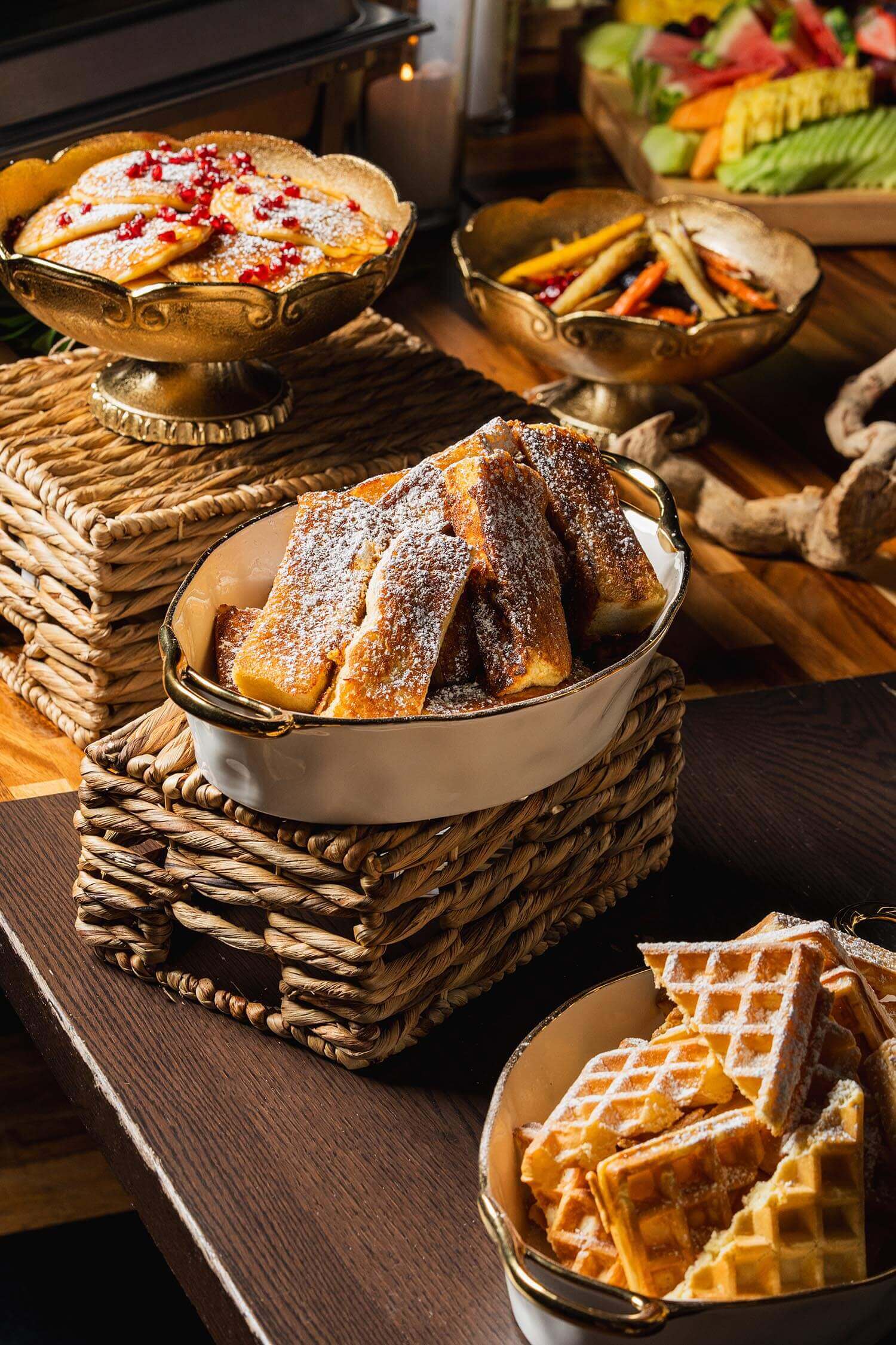 Assortment of powdered sugar-dusted pastries including waffles, French toast sticks, and pancakes served in decorative bowls on a wooden table.