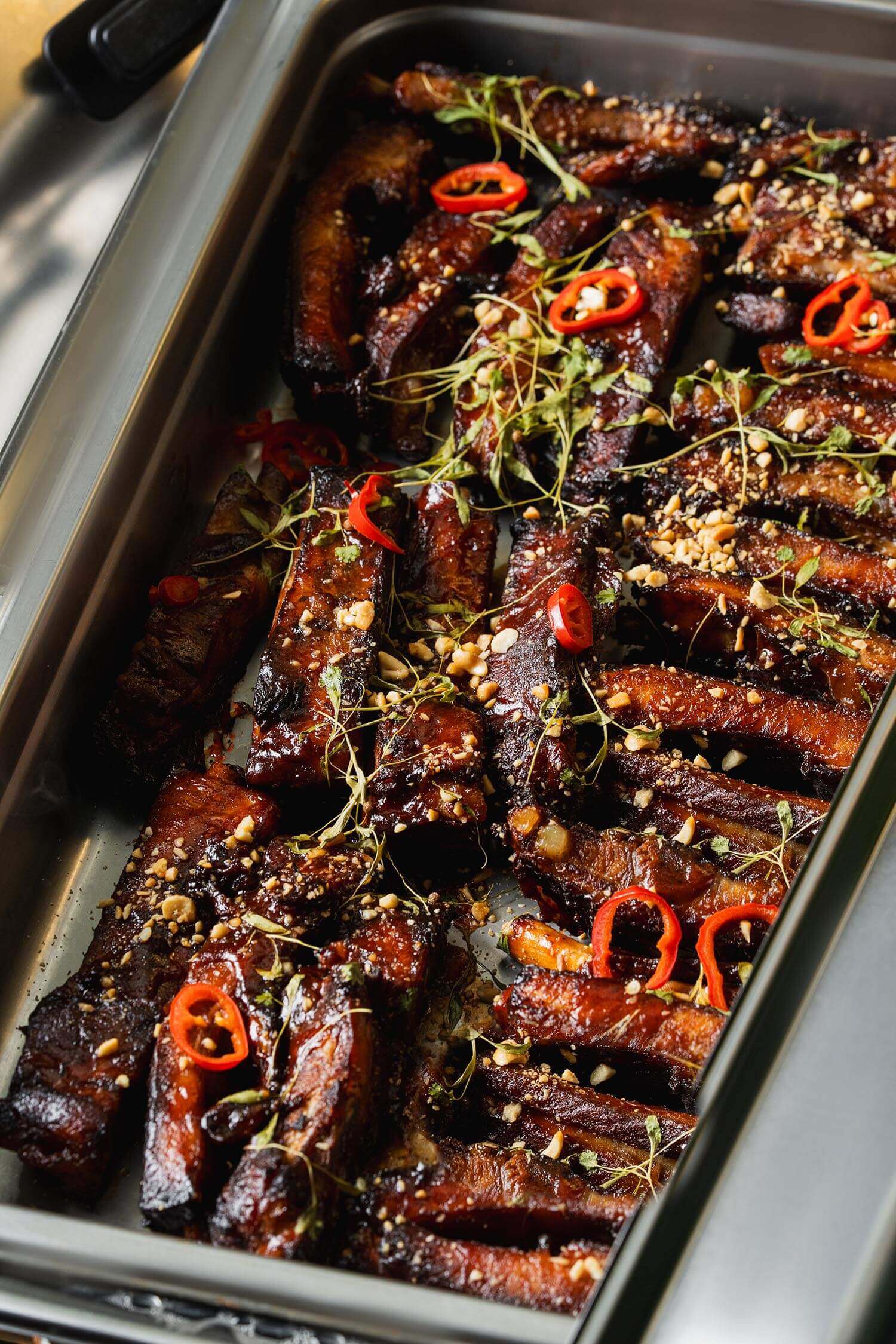 Tray of glazed cooked ribs garnished with red chili slices, fresh herbs, and chopped peanuts.