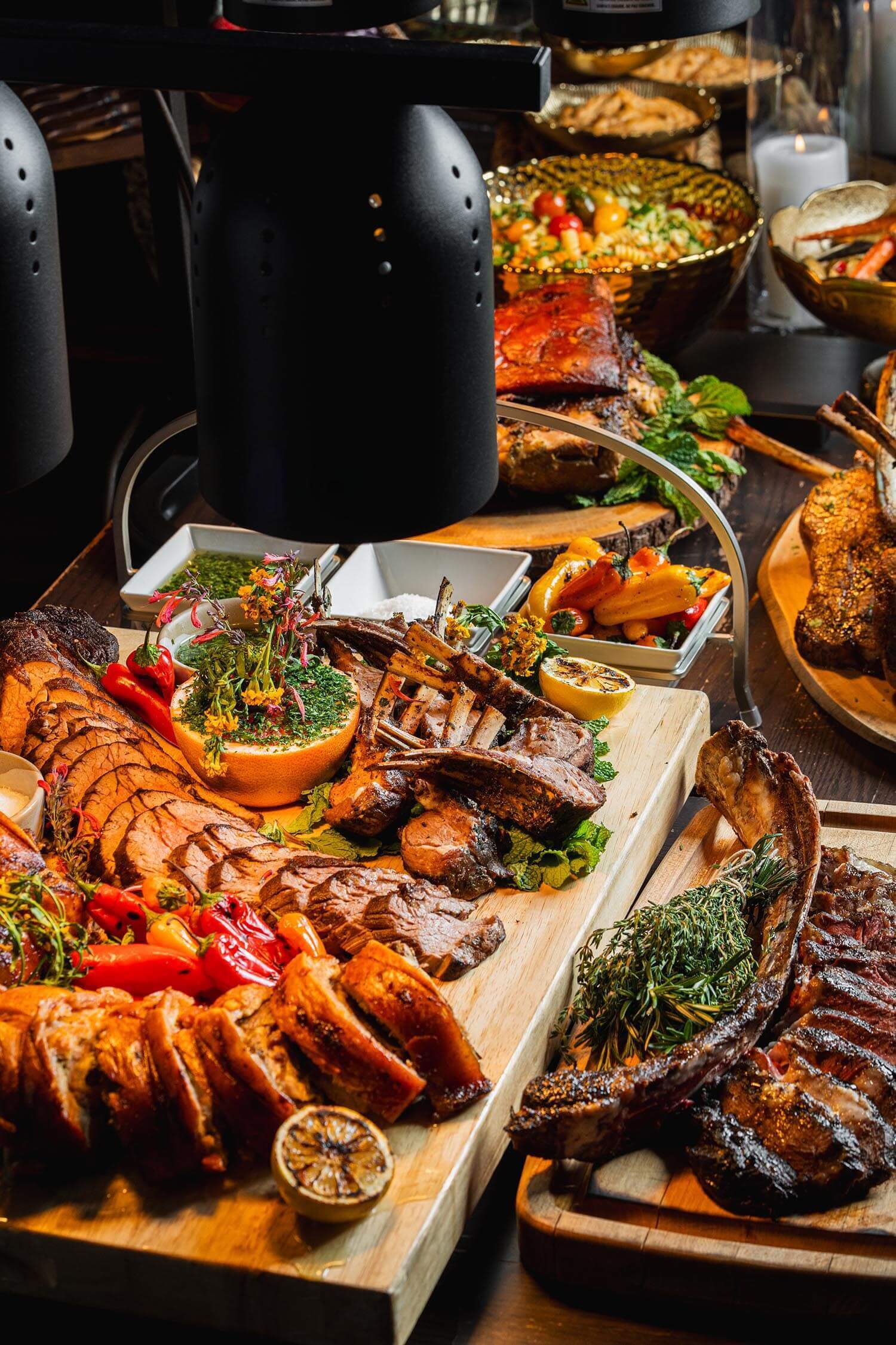 Wooden boards displaying grilled and roasted meats garnished with herbs, grilled lemon halves, and roasted peppers under warming lamps.