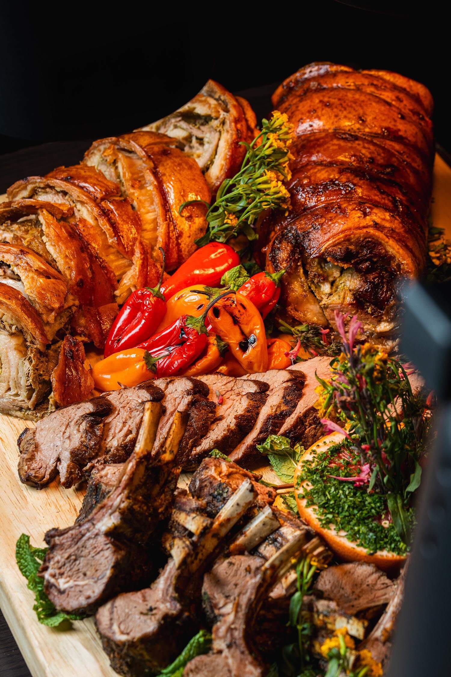 Platter with rolled roasted pork, grilled lamb chops, sliced beef, assorted colorful peppers, and garnished with herbs and greens.