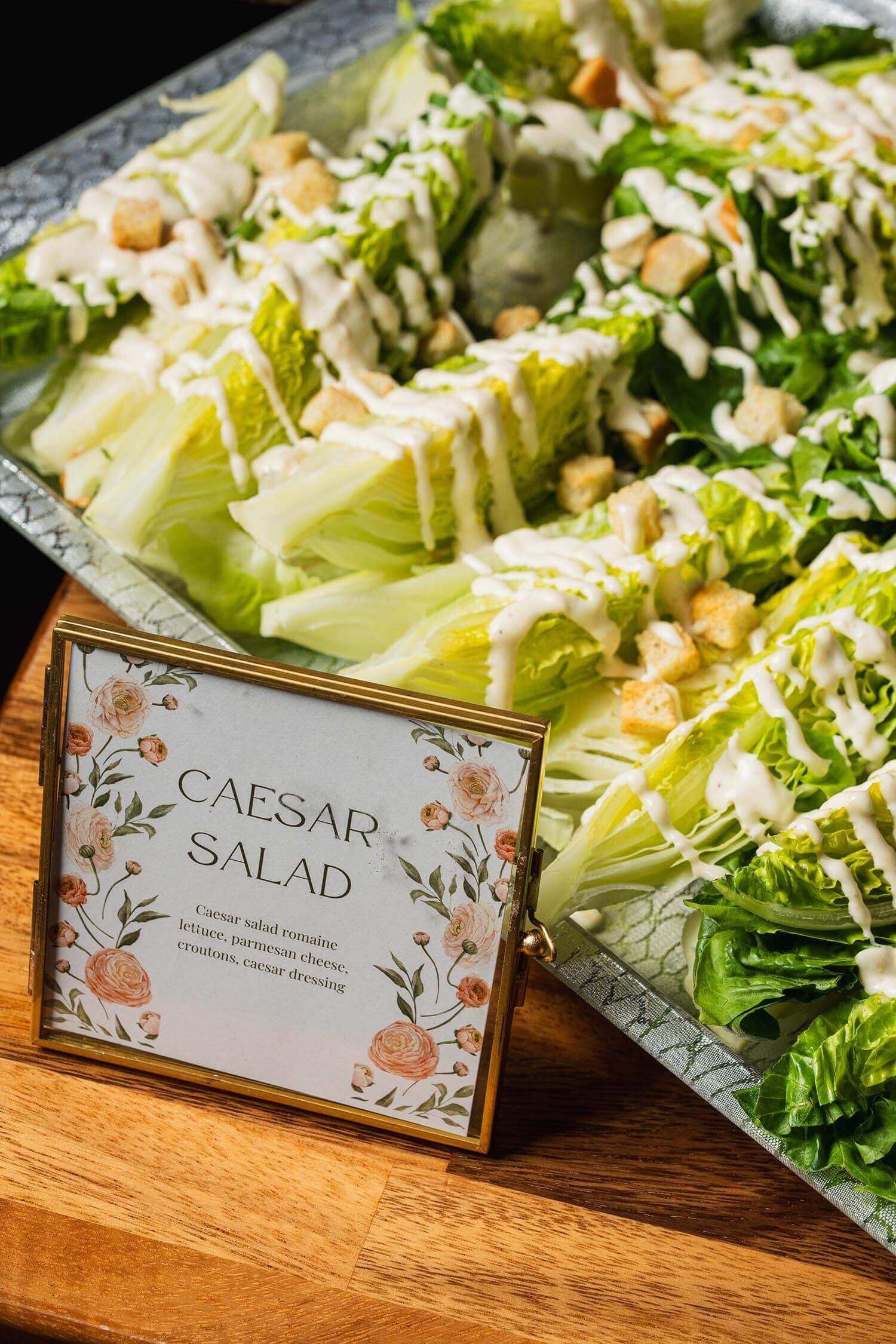 Wedge Caesar salad with romaine lettuce, croutons, parmesan cheese, and Caesar dressing on a tray with a floral-framed label.