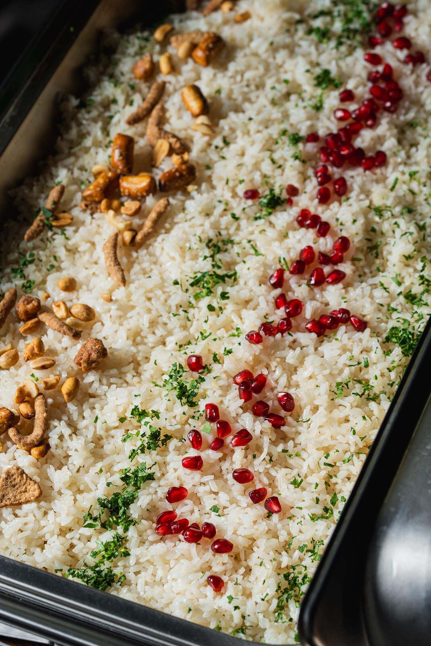 Cooked white rice garnished with chopped herbs, scattered pomegranate seeds, and mixed nuts in a black serving tray.