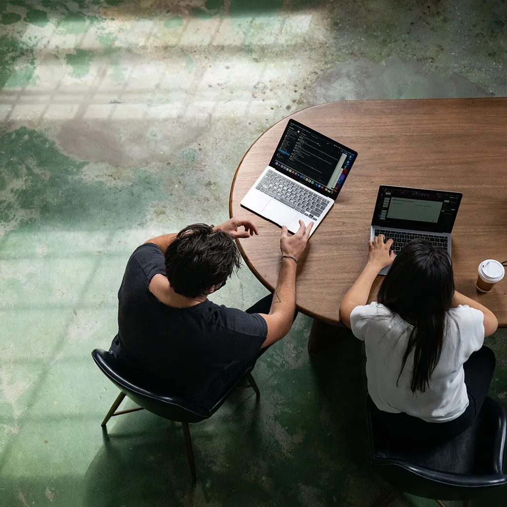 Two teammates work together on laptops at cool wooden table