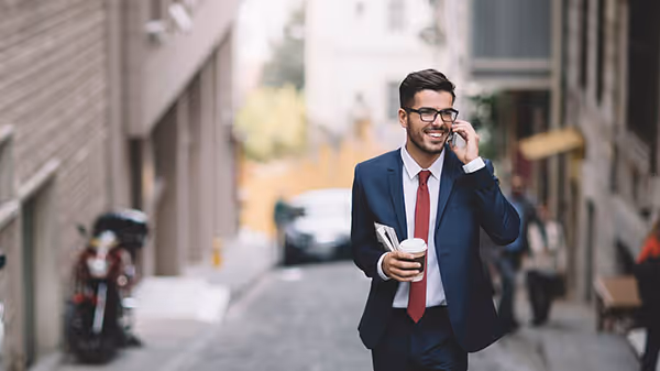 Young man in navy suit carrying a coffe cup, walking through street smiling and talking on phone