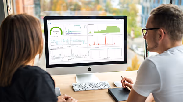 Man and woman sitting together in front of desktop monitor reviewing complex data dashboards on screen