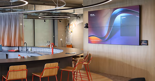 Modern office communal space with audio visual displays on wall overlooking a kitchen bar area 