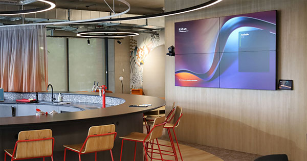 Modern office communal space with audio visual displays on wall overlooking a kitchen bar area