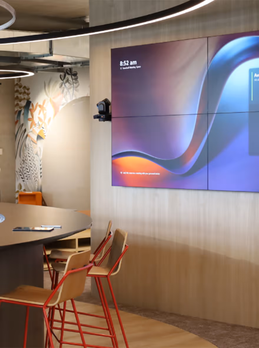 Modern office communal space with audio visual displays on wall overlooking a kitchen bar area 