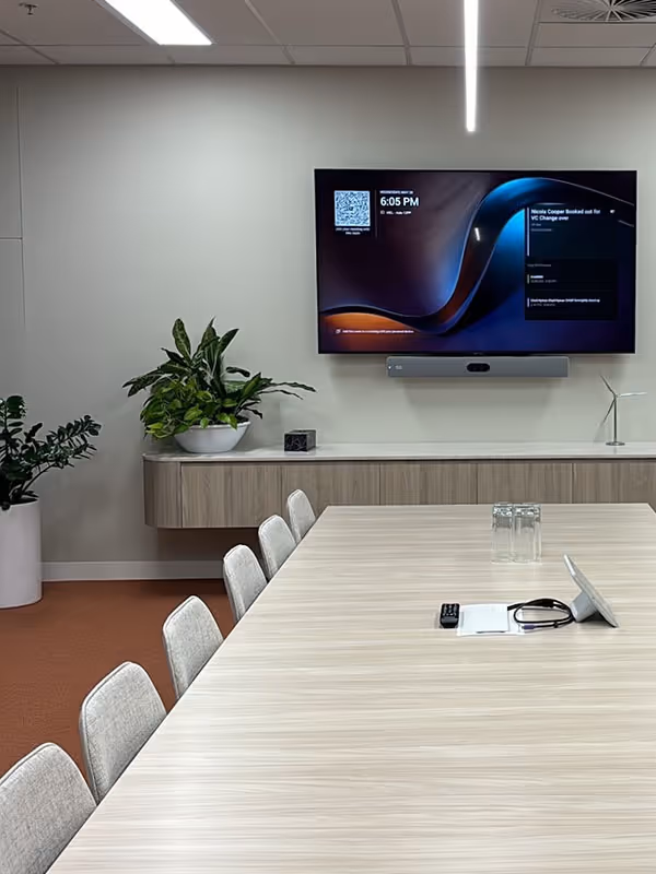 Microsoft Teams Rooms medium room in modern workplace with long linear meeting table in front of a wall-mounted display, in-built media unit and office plants.