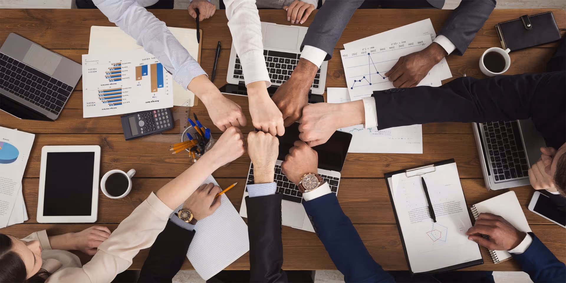 Overhead view of outstretched fists coming together in a collaborative gesture over a meeting table covered in laptops and documents