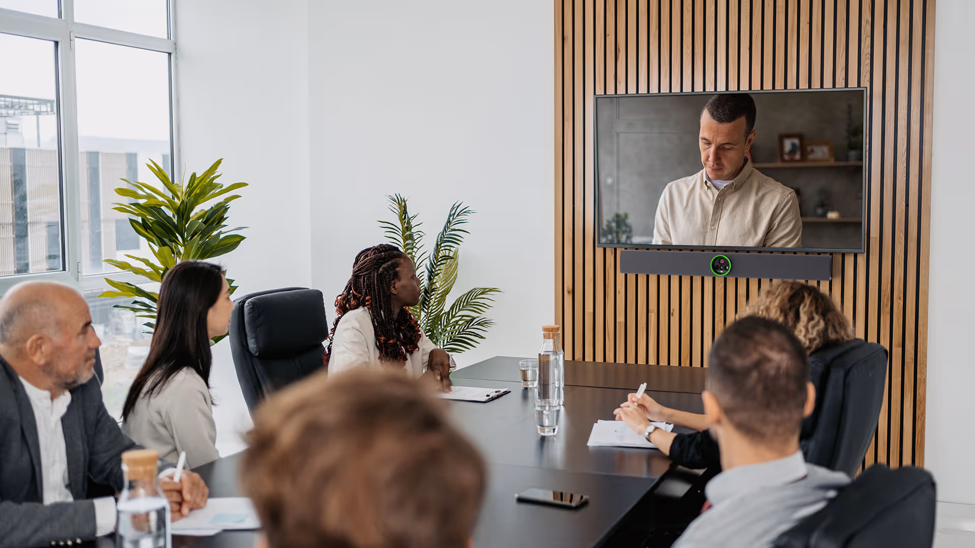 Six people in a conference room watching a man speaking on a video call displayed on a wall-mounted TV with Crestron Videobar.