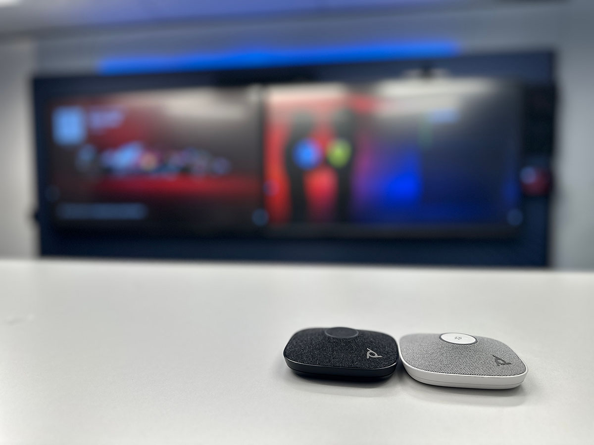 Two HP Poly Studio A2 Table Microphones, one black and one grey, placed side by side on a white meeting room table with a blurred screen in the background.