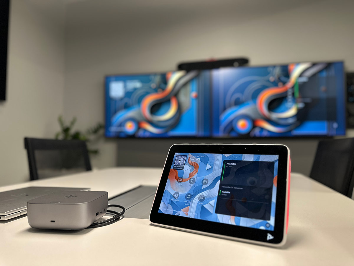 Poly TC10 Touch Controller on a meeting room table with two large monitors displaying colourful abstract art in the background.