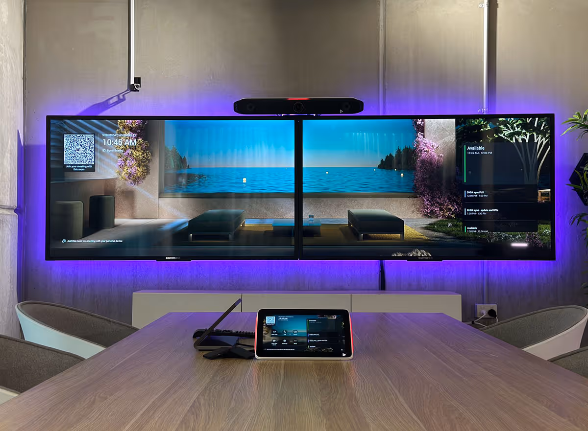 Modern conference room with dual large monitors displaying a scenic lake view and meeting schedule, a conference camera mounted above, and a control tablet on a wooden table.