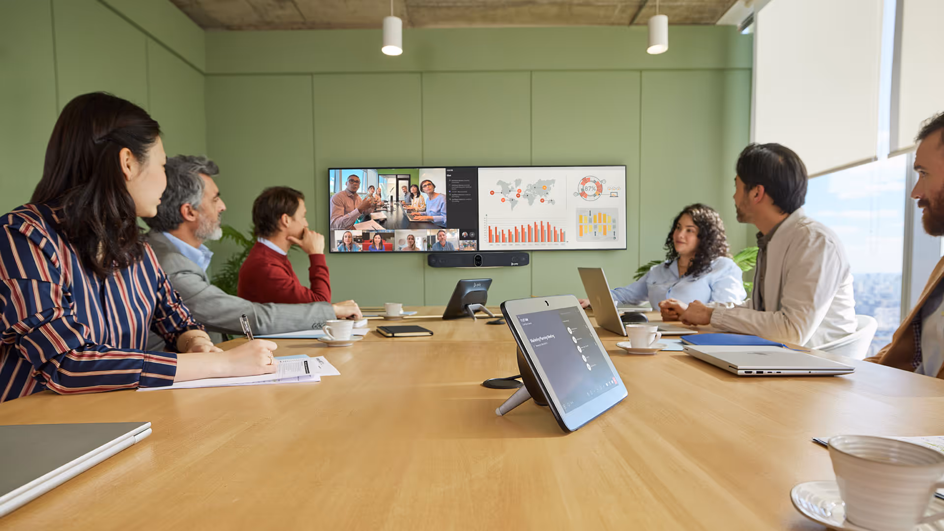 Six diverse businesspeople sitting around a conference table in a meeting room with a large screen showing video call participants and data charts.