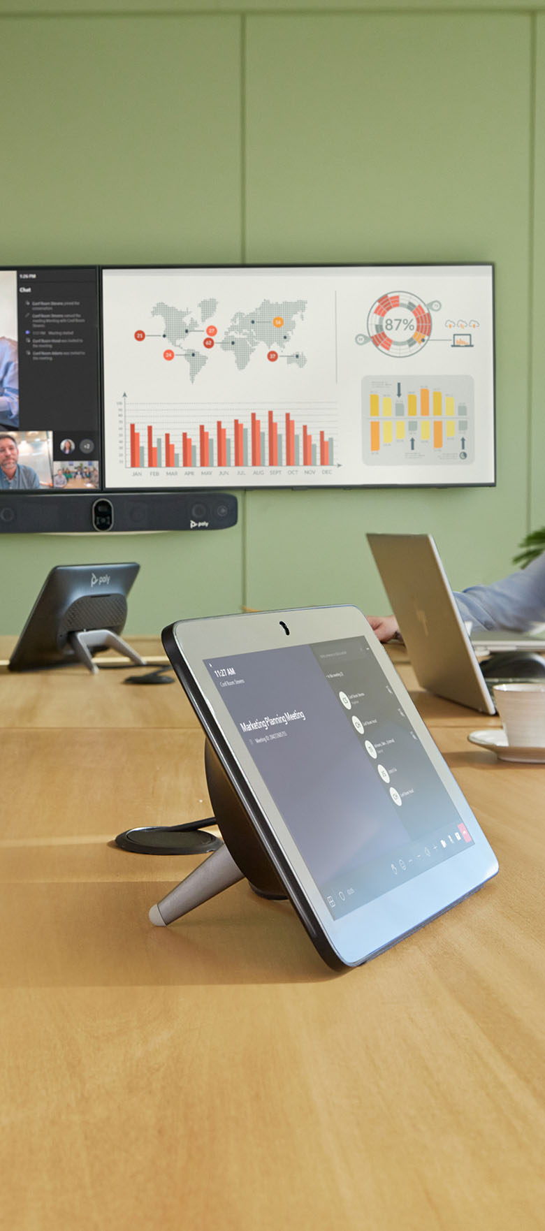 Conference room with a wooden table featuring a video conferencing device and a tablet showing a marketing planning meeting; a large wall screen displays world maps with data points, charts, and meeting participants in a video call.