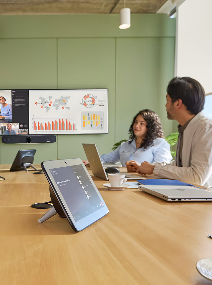 Two people seated at a conference table with laptops, viewing a screen displaying charts and a video call in a modern meeting room.