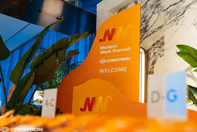 Orange signage for Modern Work Summit by Crestron with welcome message in a modern indoor space with plants.