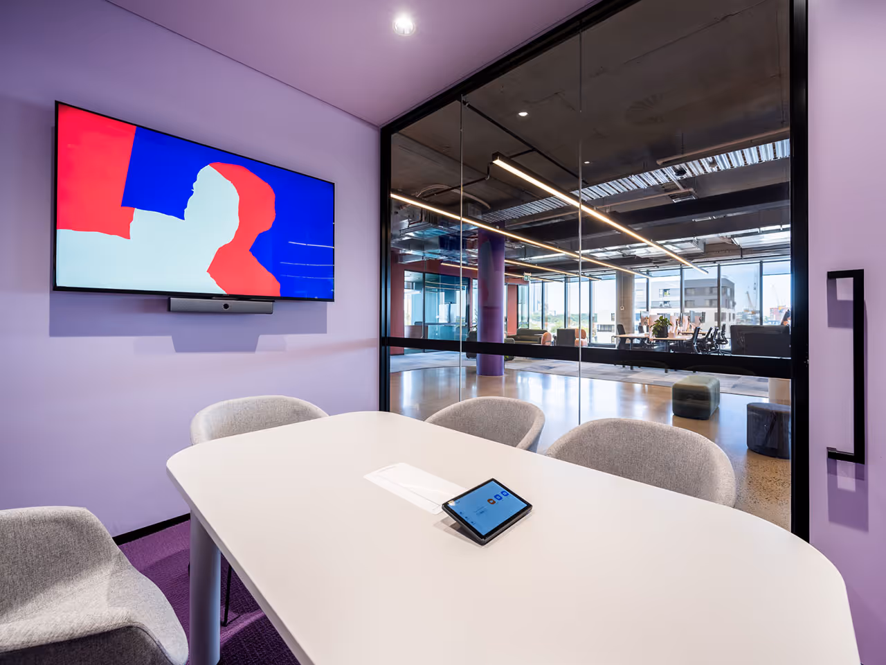 Modern small meeting room with a white table and Neat Pad, gray chairs, a wall-mounted TV with Neat Bar, and a glass wall overlooking a larger office area.
