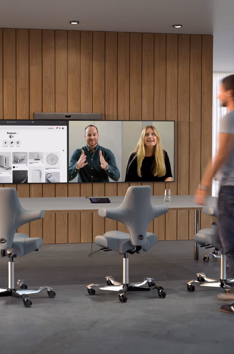 Modern conference room with three gray swivel chairs, a long table, and a wooden wall-mounted screen showing a video call with two people and a product website.