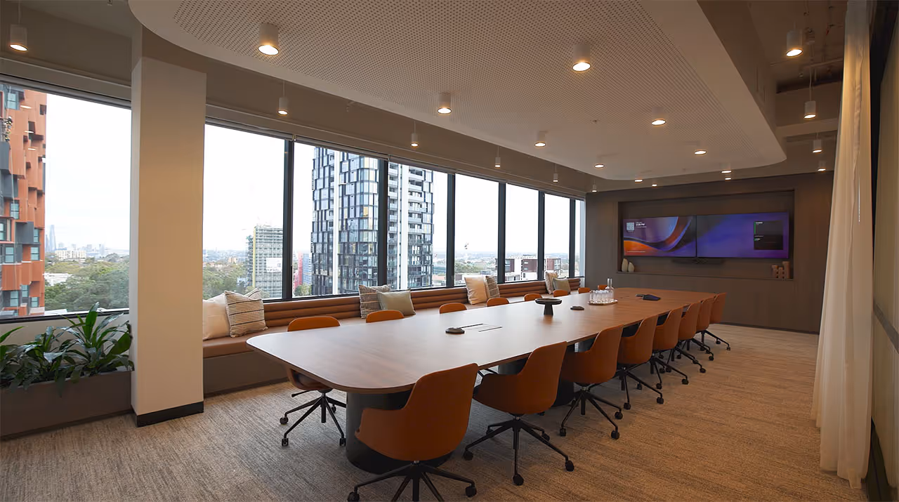 Modern boardroom with large wooden table, brown swivel chairs, built-in bench seating with pillows, and floor-to-ceiling windows overlooking city buildings.