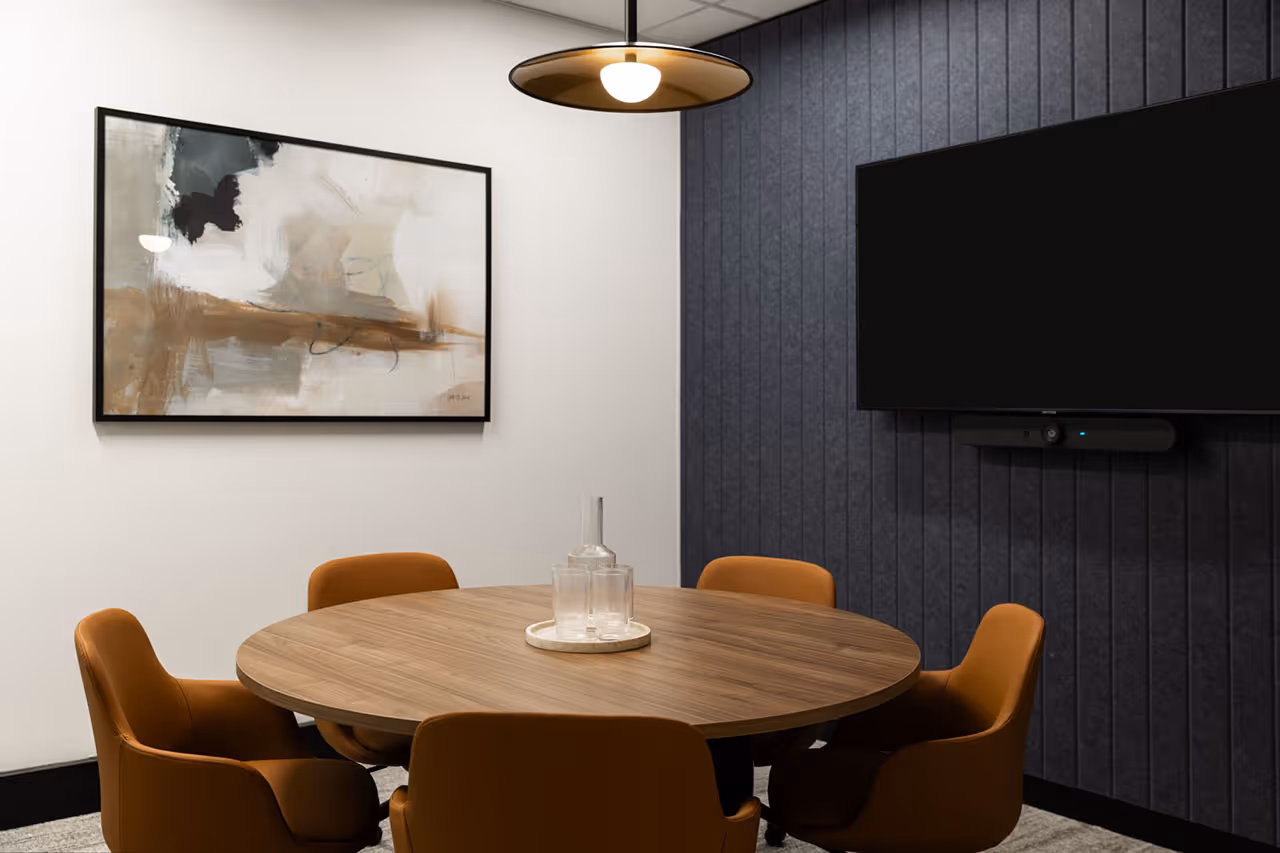 Small modern conference room with round wooden table, five brown leather chairs, abstract wall art, pendant light, and a flat-screen TV mounted on a dark paneled wall.
