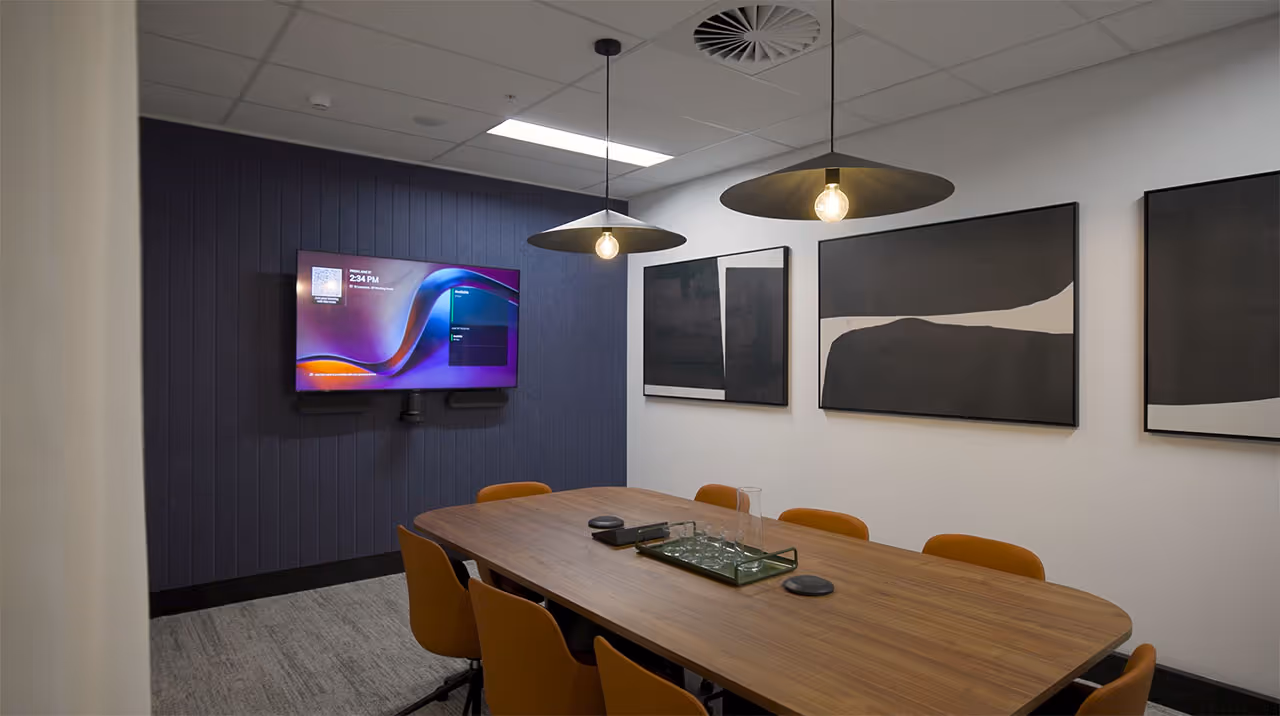 Modern conference room with wooden table surrounded by orange chairs, two pendant lights, wall-mounted screen, and abstract black and white artwork on walls.